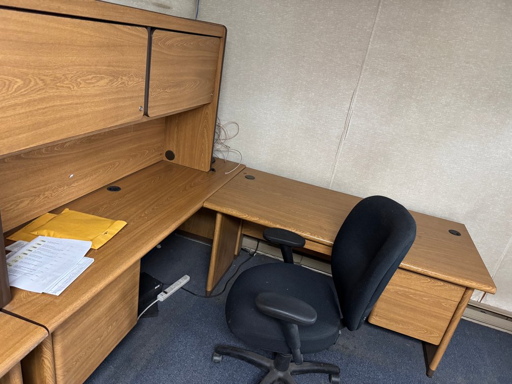 L-shaped office workstation with wood-grain desk, overhead storage hutch, drawers, and rolling office chair.