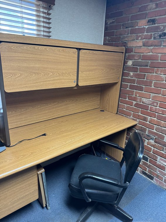 Wood-grain office workstation with overhead storage cabinets, desk surface, drawers, and rolling office chair.
