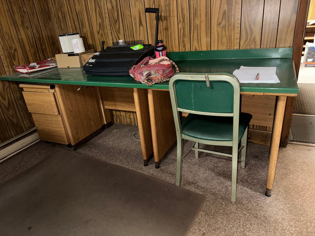 Long office worktable/desk with green top, wood-grain drawer units, metal chair, and assorted office items.