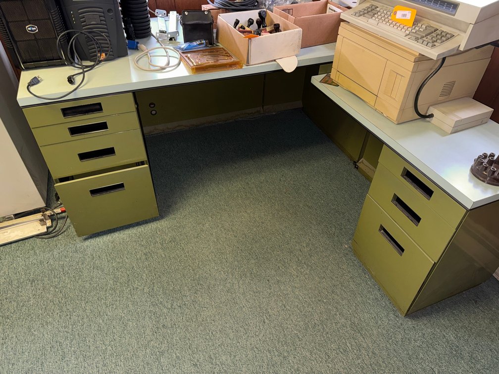 L-shaped metal office desk/workstation with multiple drawer pedestals, laminate work surface, and assorted office equipment.