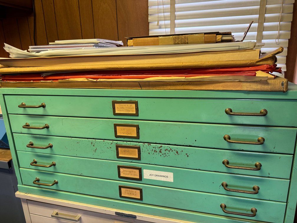 Large green flat-file/map cabinet with multiple wide drawers, label holders, and metal handles. Includes assorted maps, plans, and rolled documents stored on top.