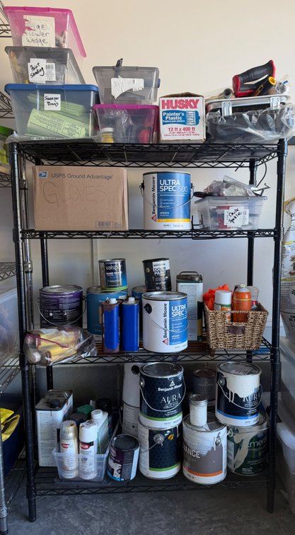 Metal shelving unit with contents including variety of different paints, sanding bars, plastic wrap, caulk, etc