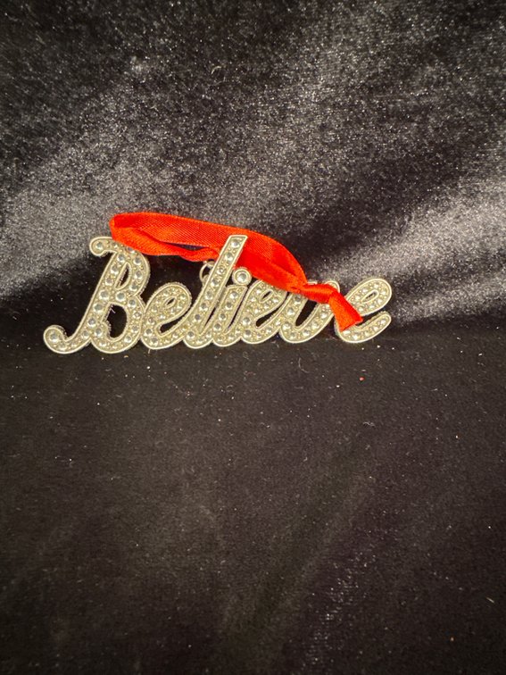 silver-plated script ornament or charm that reads "Believe," decorated with small crystal or rhinestone accents
