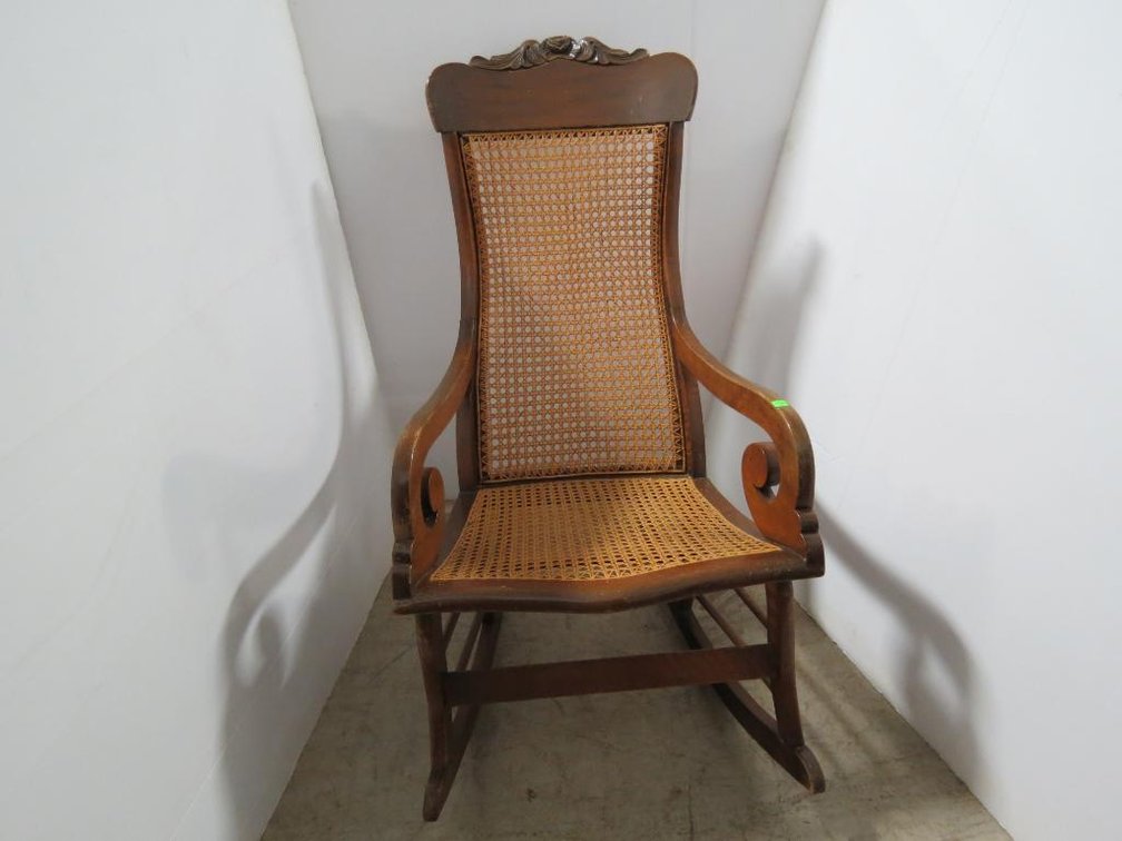 A vintage rocking chair with caned back and bottom and carving at top. Shows signs of repair.