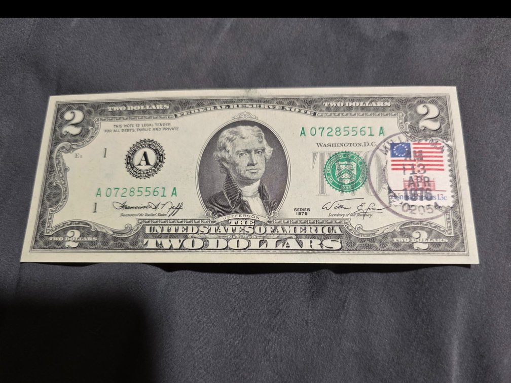Uncirculated US two-dollar Federal Reserve Note featuring a first-day-of-issue postmark and a 13-cent American Flag postage stamp.