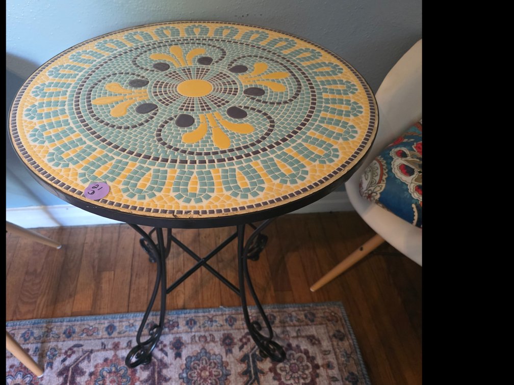 Round mosaic outdoor side table or plant stand featuring a teal, yellow, and black geometric ceramic tile top with curved black metal legs.