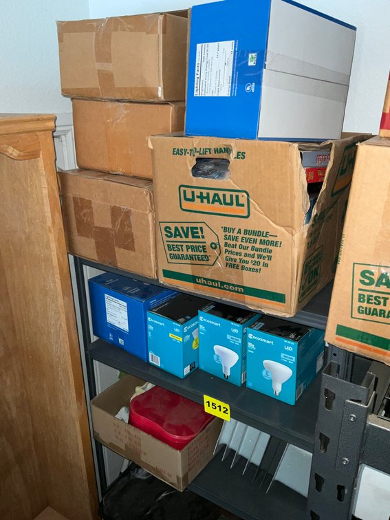 Shelving Unit And Contents In Corner Light Bulbs, VHS Movies, Plastic Kitchen Ware, Roll Of Brown Paper, White Plastic Container, Ect